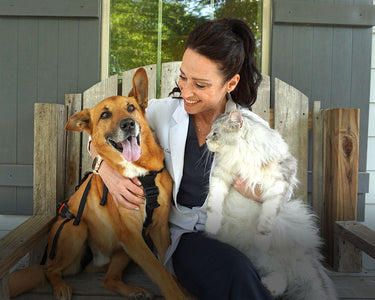Woman sitting on a wooden bench with a dog and a cat, smiling outdoors.